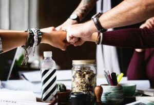 A diverse group of people fist-bumping over a desk, showing teamwork, unity, and collaboration in a casual work setting with snacks, drinks, and stationery on the table.