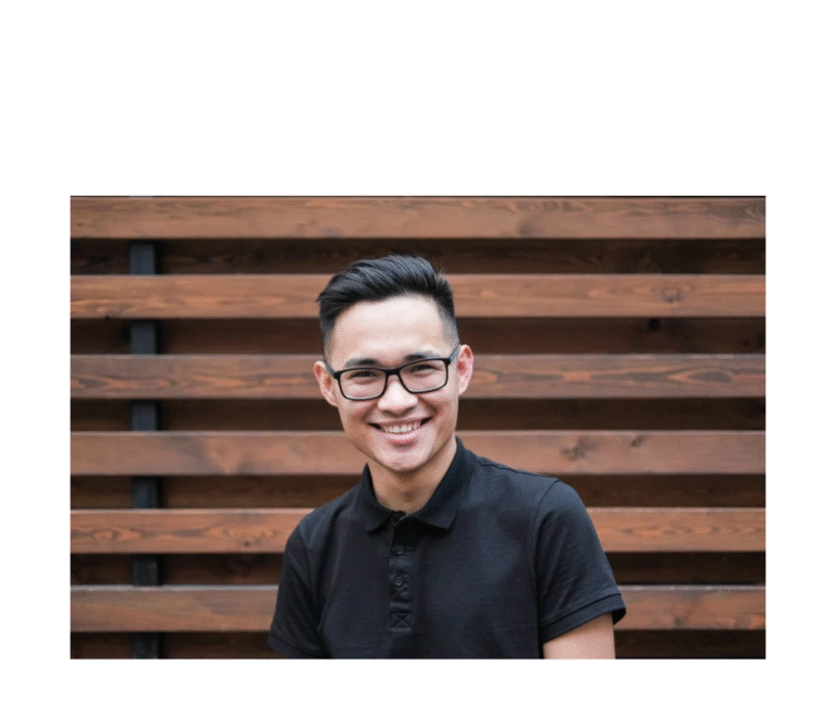 A man wearing glasses and a black shirt standing in front of a wooden wall, smiling